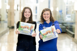 Two women wearing black (left) and blue (right) holding a copy of the book "Sam Becomes a Sycamore."