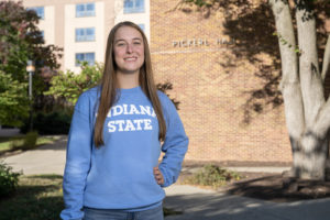 Woman with long brown hair wearing a blue Indiana State sweatshirt, poses in front of Pickerl Hall.