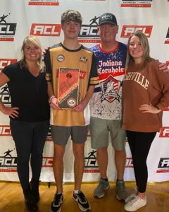 Left to right: Woman with shoulder-length blonde hair wearing a black T-shirt and dark jeans; Man with brown hair wearing a gold and black athletic shirt and gray short holds ACL Champion award; Man in Indiana Cornhole T-shirt and gray shorts wearing a MOPO baseball cap; Woman with shoulder-length blonde hair wearing a dark orange Nashville sweatshirt and black pants.