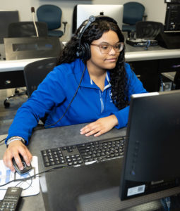 Jabrea Jones, an African American woman, wears a headset and is working on a computer while wearing a long-sleeve blue ISU Student Media jacket.