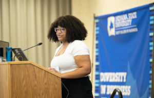 Jabrea Jones, an African American woman, poses at a podium while wearing a white dress shirt and black dress pants.