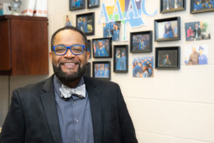 Kale Walker, a middle-aged African American male, poses in front of a wall with framed pictures and an AAAC logo. He wears blue glasses and a blue and white bow tie.
