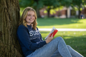 Adalyn McMurray, a young white woman with shoulder-length wavy blond hair, sits next to a tree. She wears a long-sleeve blue STATE Honors College T-shirt, blue jeans, and she's holding a red book.