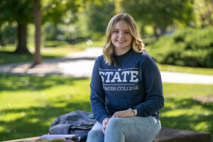 Adalyn McMurray, a young white woman with shoulder-length wavy blond hair, sits on a bench. She wears a long-sleeve blue STATE Honors College T-shirt and blue jeans. A gray backpack is next to her.