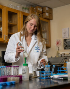 Elynor Head, a young white woman with dark straight blond hair, poses with lab equipment in a lab. She wears a white lab coat.