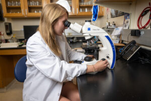 Elynor Head, a young white woman with dark straight blond hair, is looking in a microscope in a lab. She wears a white lab coat.