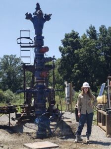 Elynor Head, a young white woman with dark straight blond hair, poses next to drilling machinery at a drill site. She wears a long-sleeve tan shirt, dark blue pants, and a white hard hat.