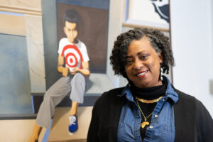 Valerie Craig, an African American woman, poses in front of a painting of a young child wearing a white T-shirt with a red bullseye. Craig wears a blue jean top, a black jacket, and gold necklaces.
