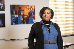 Valerie Craig, an African American woman, poses in front of a wall and window. The wall has photographs of Indiana State students at graduation. Craig wears a blue jean top, a black jacket, and gold necklaces.