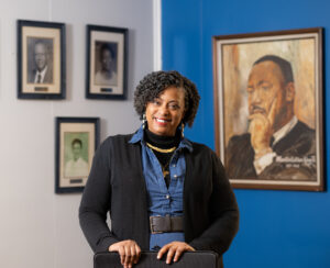 Valerie Craig, an African American, poses in front of a wall of photographs and a painting of Dr. Martin Luther King, Jr. She wears a blue jean dress, a black jacket, and gold necklaces.