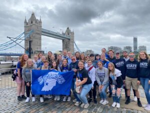A group of young adults posing in front of the London Bridge, holding a blue Sycamores flag. 