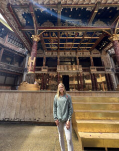 Elisabeth Kerby, a young white woman with long blonde hair, poses at Shakespeare's Globe Theatre. She wears white pants and a light green shirt.