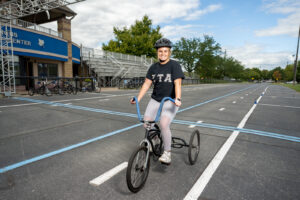 Elisabeth Kerby, a young white woman, poses on a tricycle on a race track. She wears white athletic leggings and a black T-shirt with ZTA letterings. 