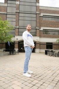 Gage Fraser, a young white man with light brunette hair and a beard and mustache, poses in front of a brick building. He wears a white sweatshirt with Indiana State blue lettering.