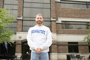 Gage Fraser, a young white man with light brunette hair and a beard and mustache, poses in front of a brick building. He wears a white sweatshirt with Indiana State blue lettering.