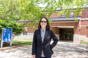 Katherine Lee, a Korean-American woman with shoulder-length straight black hair, poses between two brick posts, surrounded by trees. She wears a navy blue dress coat, a blue-and-white striped dress shirt, and dark sunglasses.
