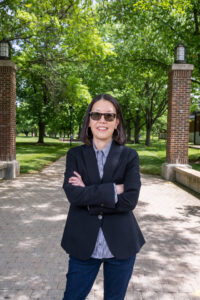 Katherine Lee, a Korean-American woman with shoulder-length straight black hair, poses between two brick posts, surrounded by trees. She wears a navy blue dress coat, a blue-and-white striped dress shirt, and dark sunglasses.