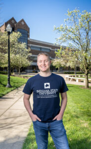 Kohler Kerber, a young white man with short blonde hair, poses in front of a brick academic building and trees. He wears a blue T-shirt with Indiana State University Bailey College of Engineering & Technology printed on the front.