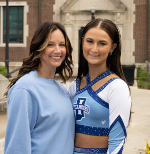 Macy Ramey, a young white woman with straight long brunette hair, poses next to Angie Ramey. She wears a blue and white cheerleading uniform. Angie, a middle-aged white woman with wavy brunette hair, wears a blue sweatshirt. They pose in front of a brick building.