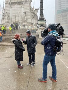 On the left, Taylor Schaffer is being interviewed by a man while a cameraman is to the right. All three individuals wear dark winter coats and they're standing outside in the snow near a monument.