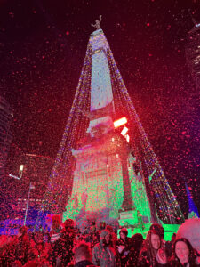 At night, a lights display at a tall monument with strings of lights. A crowd of individuals are below the monument.