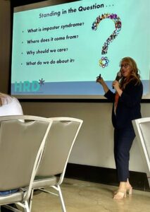 Andrea Butcher, a woman with wavy blonde hair, is giving a presentation. A PowerPoint screen is behind her with a giant question mark and writing about Imposter Syndrome. She wears dark dress pants and a matching blazer. Chairs are in front of her.