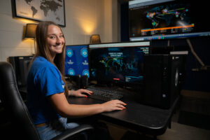 Payton Vallee, a female student with long straight brown hair, poses in front of a desktop computer with a digital world map and "Fireeye Cyber Threat Map" on the screen. She wears a blue Indiana State T-shirt.