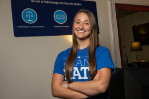 Payton Vallee, a female student with long straight brown hair, poses in front of a blue sign for the School of Criminology and Security Studies. Her arms are crossed and she wears a blue Indiana State T-shirt.