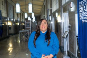 Courtney Richey-Chipol, a woman with long black hair, poses in a grey hallway. She wears a blue turtleneck sweater. A banner reading "West Central Indiana Small Business Development Center" is on the wall behind her.