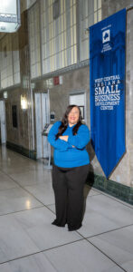 Courtney Richey-Chipol, a woman with long black hair, poses with her arms crossed in a grey hallway. She wears a blue turtleneck sweater. A banner reading "West Central Indiana Small Business Development Center" is on the wall behind her.