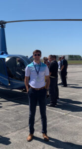 Dylan Maupin, a white male student with short brown hair, poses in front of a helicopter. A group of men are behind him. Maupin wears sunglasses, a white-and-blue polo shirt, and dark pants.