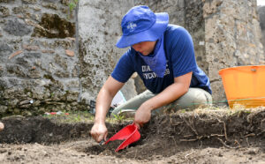 A student wearing a blue Indiana State Sycamores T-shirt, a blue hat with the Sycamores logo, and a blue bandana. The student is digging in soil with a stone structure in the background.
