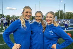 Three female students with blonde hair in ponytails pose on a football field. They wear blue Spirit Squad jackets.