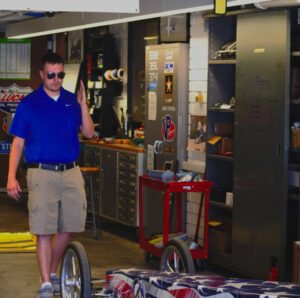 A white male student with short brown hair is standing in front of a patriotic-themed dragster. He wears dark sunglasses, a blue polo shirt with a white undershirt, and khaki shorts. His left hand is raised as he looks at the dragster. Metal cabinets and tools are visible in the background. 