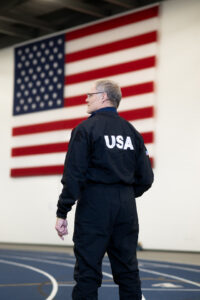 A white male with short grey hair poses in front of an American flag with his back facing the camera. He wears a black athletic jumpsuit with USA in white lettering, and he holds a red helmet against his right hip.