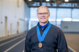 A white male with short grey hair stands on an indoor racing track. He wears a black athletic jumpsuit and he wears a medal around his neck.