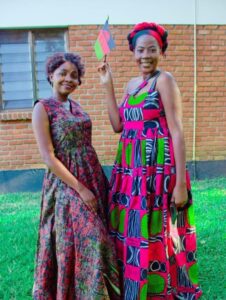 Two Black women wearing traditional African attire, posing in front of a brick building. The woman on the right holds a black, red, and green flag.
