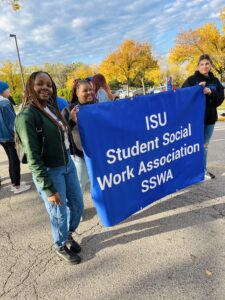 On the left is a Black woman with light brown dreadlocks participating in a walk. She holds a blue "ISU Student Social Work Association SSWA" banner with other students. 