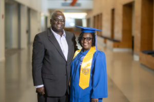 On the left is a middle-aged Black male wearing a dark grey suit with a white undershirt. To his right is a middle-aged Black woman with shoulder-length black hair. She wears a blue graduation gown, a blue graduation cap, and a yellow graduation sash. They stand in a hallway. 
