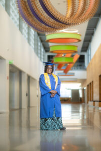 A middle-aged Black woman with shoulder-length black hair standing in a colorful hallway. She wears a blue graduation gown, a blue graduation cap, and a yellow graduation sash.