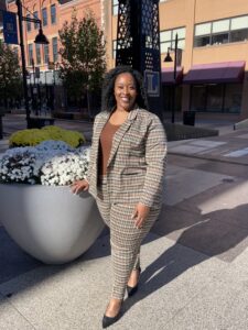 Bre'Anna Donaldson, a Black woman, stands outside next to a potted plant. She wears a tan checkered pantsuit with a brown tank top shirt. Buildings are in the background.