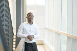 A Black male student wearing a white dress shirt poses in a hallway next to a railing.