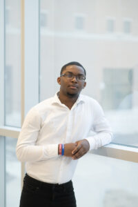 A Black male student wearing a white dress shirt poses in front of glass windows. 