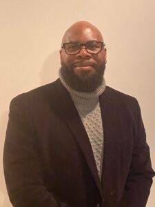 Dr. Marcus McChristian, a Black man, wears a black suit jacket and a grey turtleneck sweater underneath the jacket. He poses for a photo.