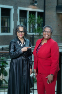 On the left, Dr. Kandace Hinton, wears a black trench coat. On the right, Dr. Mary Howard-Hamilton, wears a red pantsuit. Hinton points to Howard-Hamilton. They stand in front of a railing in the atrium of University Hall in the Bayh College of Education.