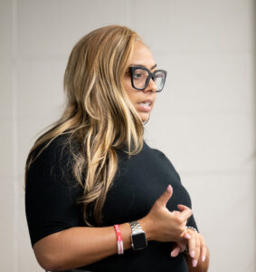 Andrea Brown, a Black woman with long light brown hair, stands in a classroom setting. She wears black glasses, a black turtleneck sweater, and grey plaid dress pants.