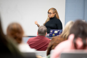 Andrea Brown, a Black woman with long light brown hair, stands in a classroom setting in front of a whiteboard. She wears black glasses, a black turtleneck sweater, and grey plaid dress pants. Other students are blurred, sitting in front of her.