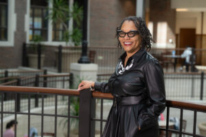 Dr. Kandace Hinton, a Black woman with short black dreadlocks, poses in an atrium, leaning against a brown railing with her right hand on the railing. She wears black glasses, a black-and-white necklace, and a black leather jacket dress.