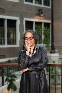 Dr. Kandace Hinton, a Black woman with short black dreadlocks, poses in an atrium, leaning against a brown railing with her right index finger on the side of her face. She wears black glasses, a black-and-white necklace, and a black leather jacket dress.