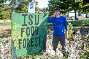 A white male student with short brown hair stands in a garden with a large green sign reading "ISU FOOD FOREST" to his left. He wears a blue long-sleeved shirt with a white sycamore leaf on the front, and grey pants.